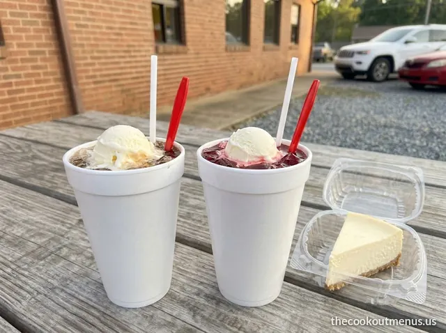 Two soda floats with vanilla soft serve in foam cups next to a slice of New York-style cheesecake